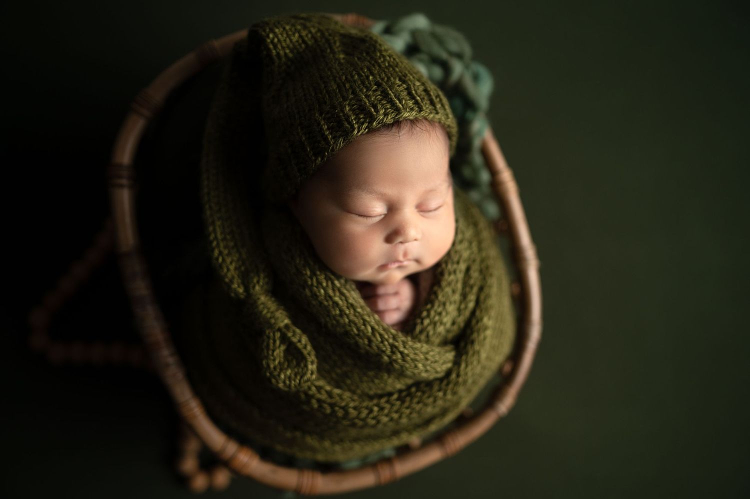 Newborn cozily wrapped in olive green knit blanket in rustic wooden bowl.