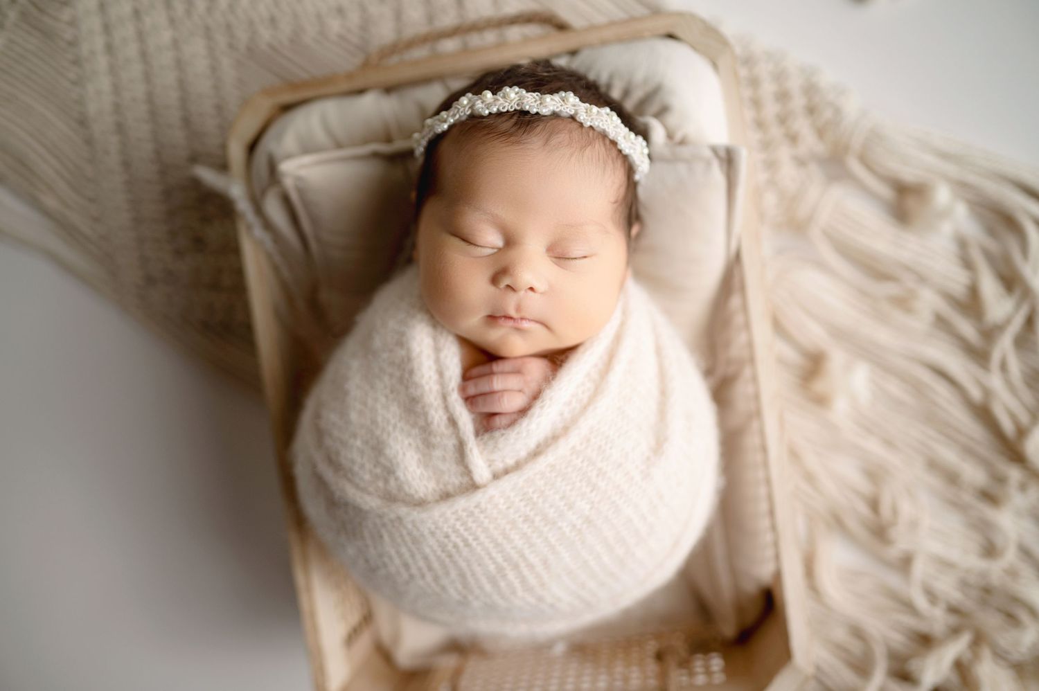 Tiny newborn wrapped in white swaddle on cream textured backdrop with soft studio lighting.