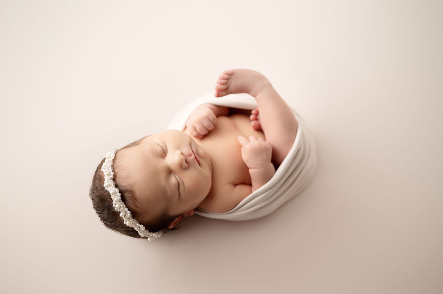 Sleeping newborn baby wrapped in white fabric with delicate lace headband on beige backdrop.