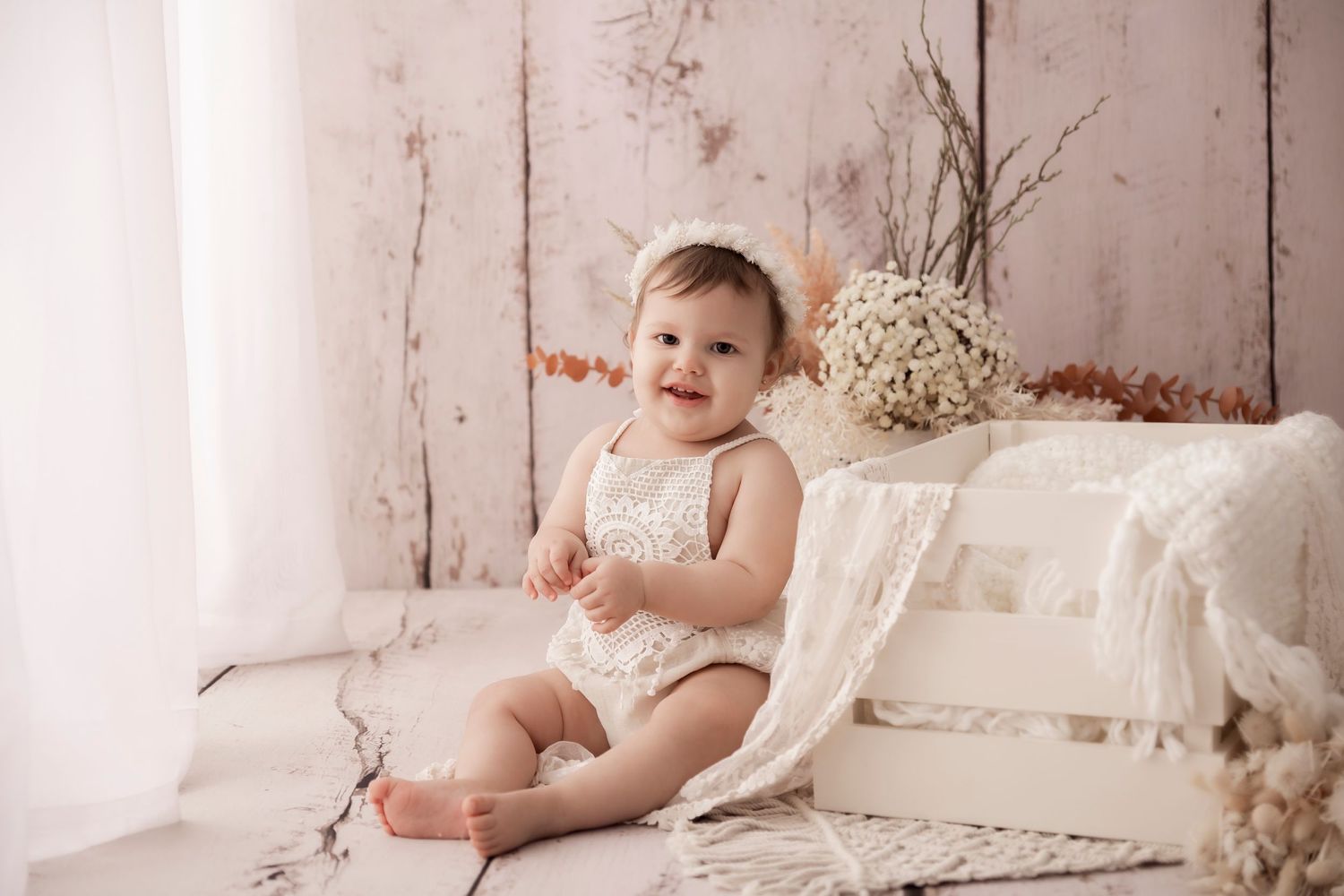 Baby in white romper sits on textured blanket against rustic wooden backdrop with dried floral decorations. Townsville, QLD, Australia