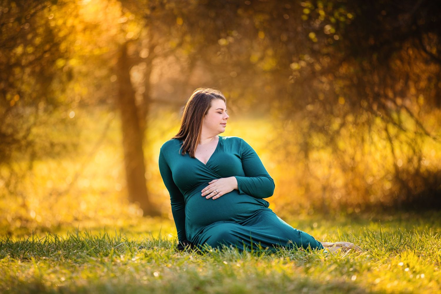 Expectant mother in teal dress sits peacefully on grass during golden sunset.