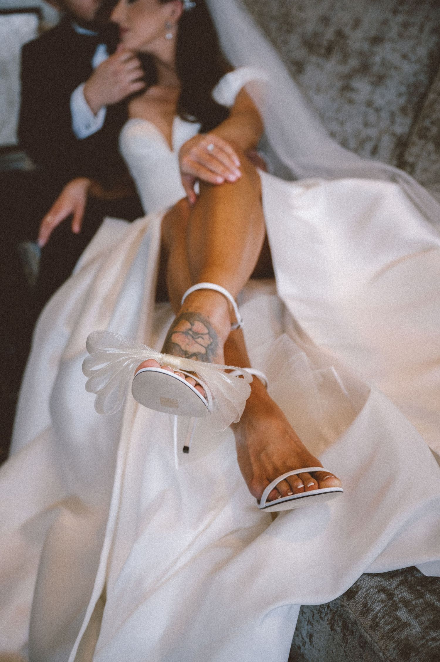 Close-up of a bride's leg showing white wedding shoes and a small tattoo against flowing white dress fabric.