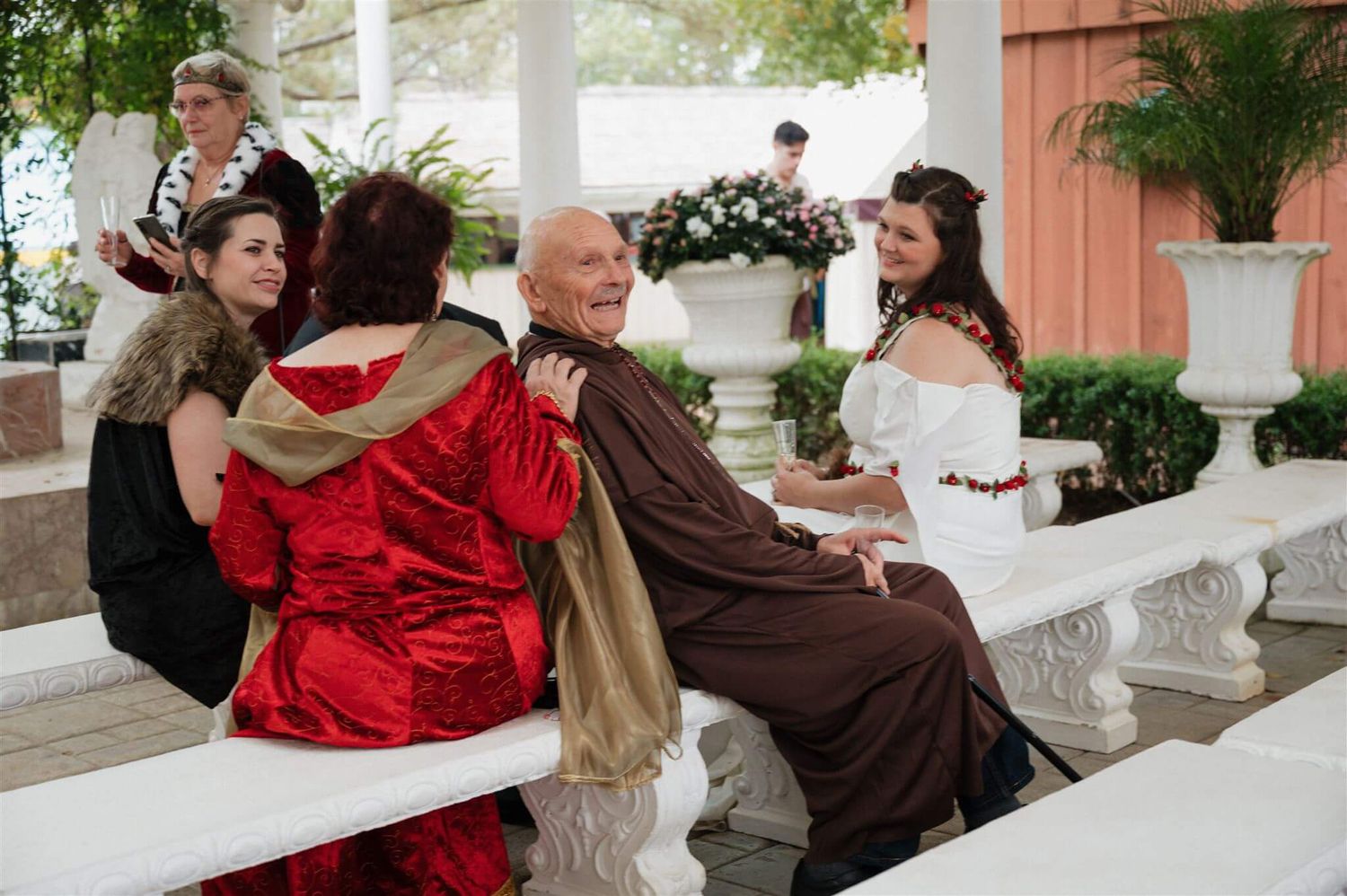 Wedding party in renaissance costumes relaxing on white bench during outdoor celebration.