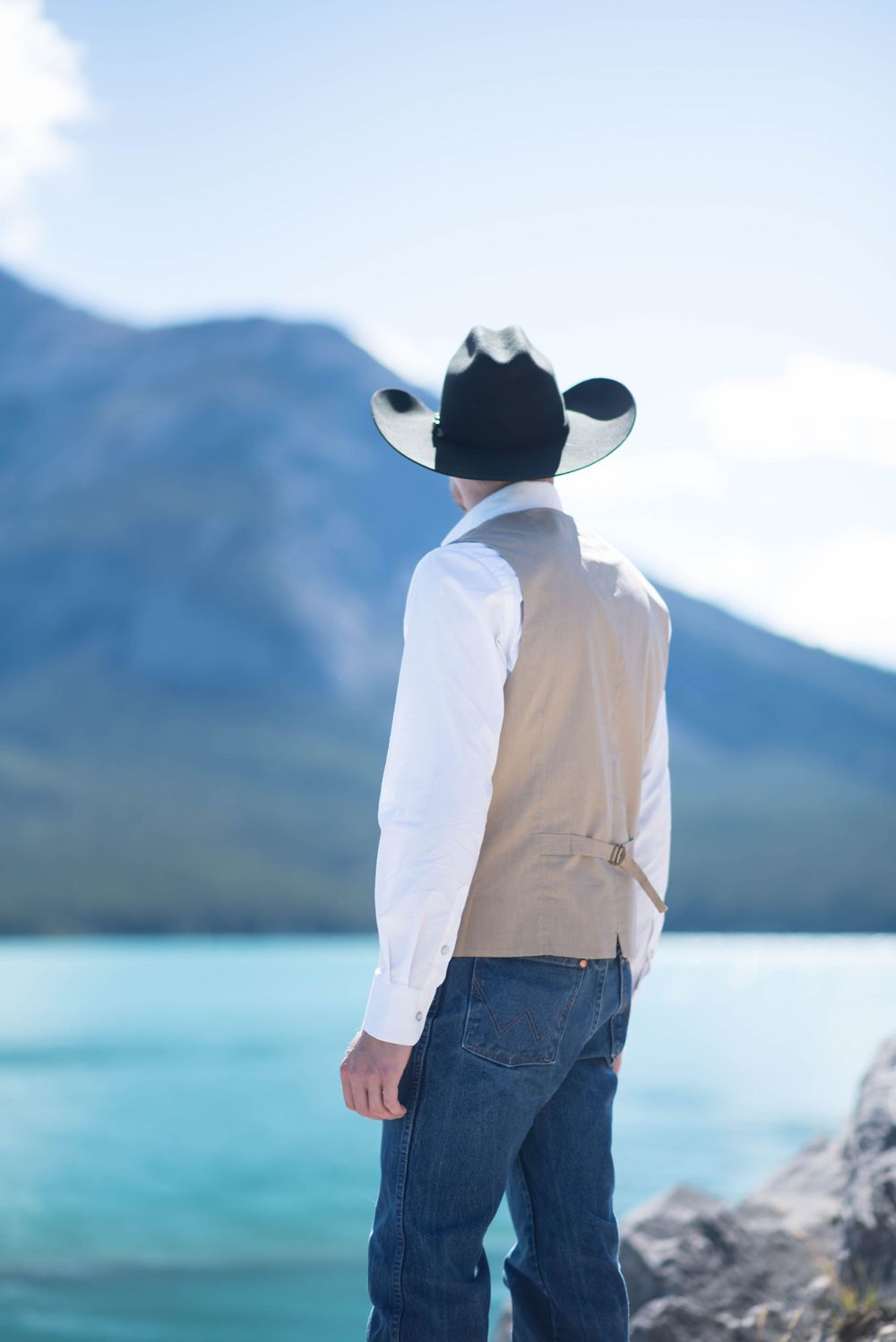 Cowboy in white shirt and hat looking out at turquoise mountain lake.