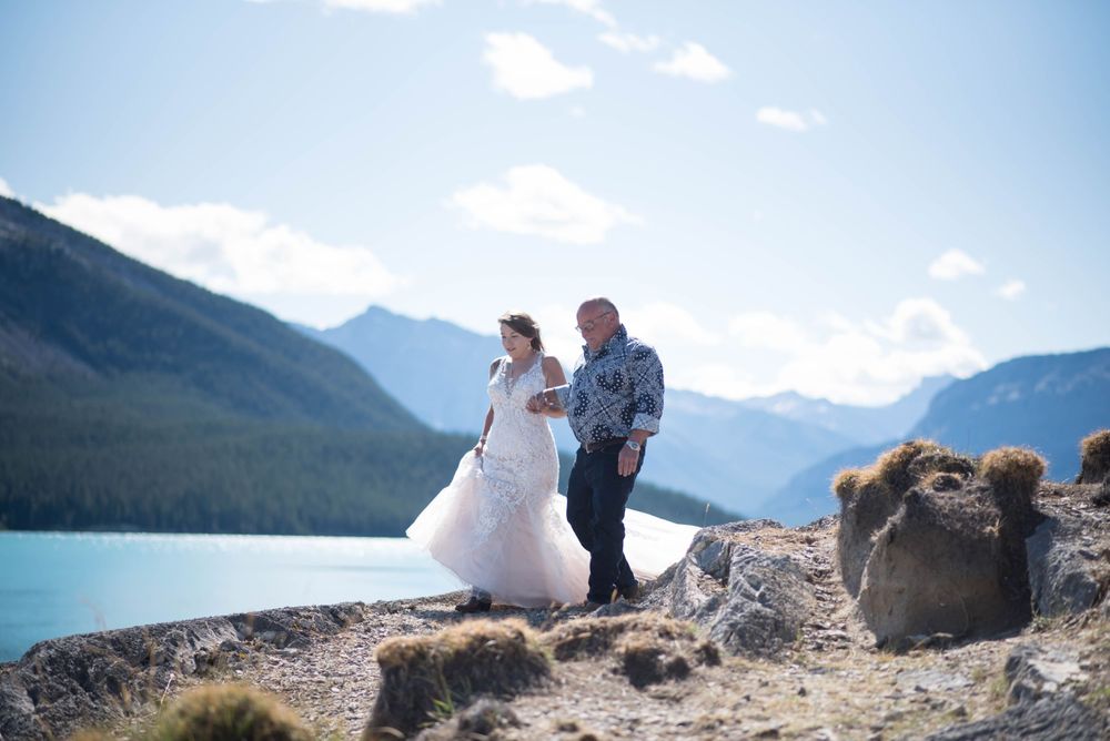 A wedding ceremony takes place on a rocky overlook with mountain vistas and blue lake below.