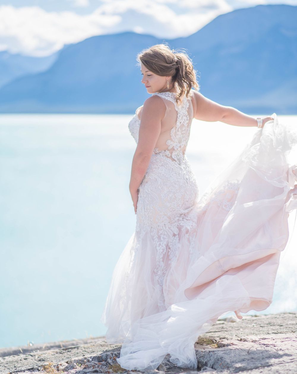 Lace wedding dress flows in breeze against spectacular mountain lake backdrop.