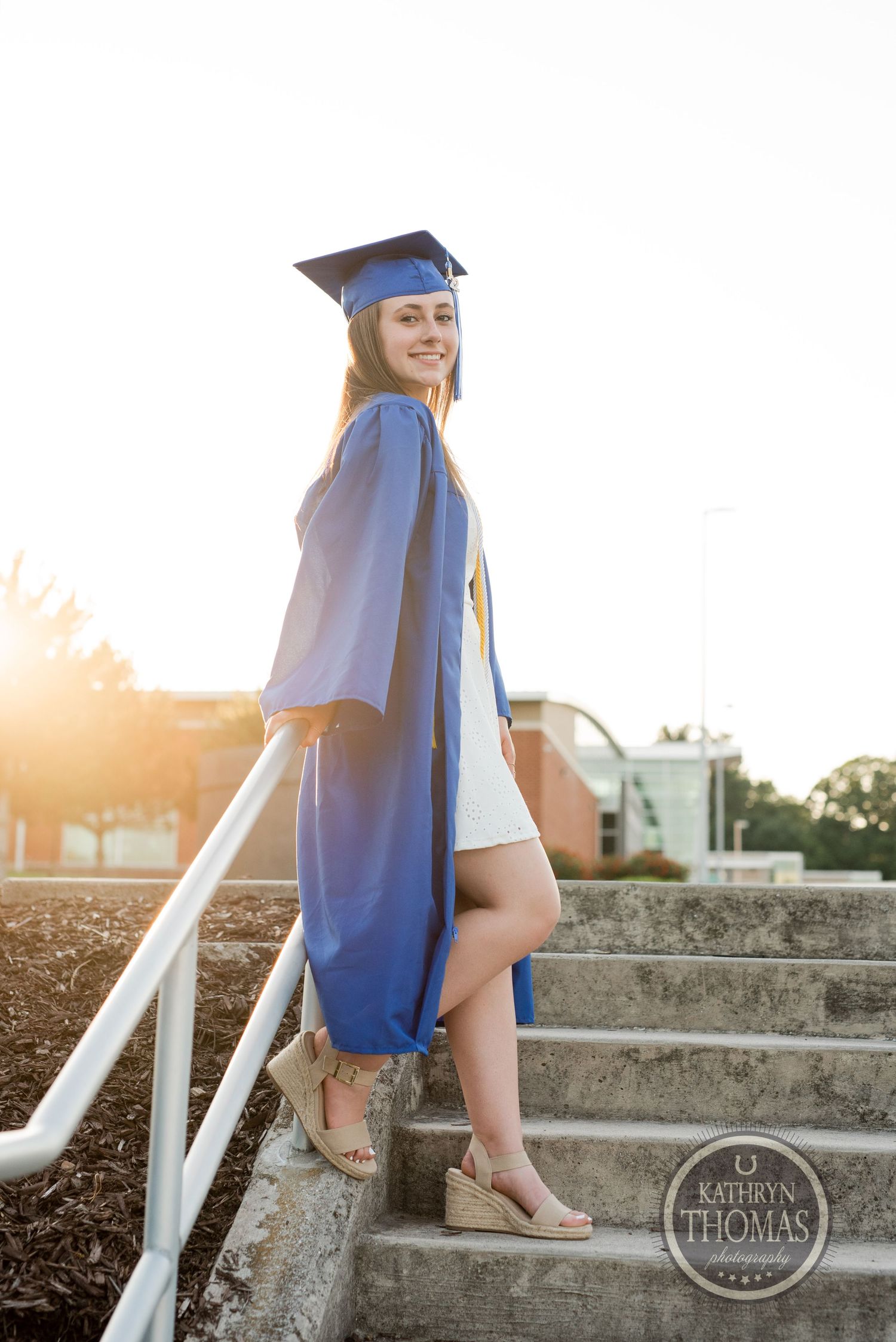 Graduate poses on outdoor steps at sunset in blue graduation gown and white dress.