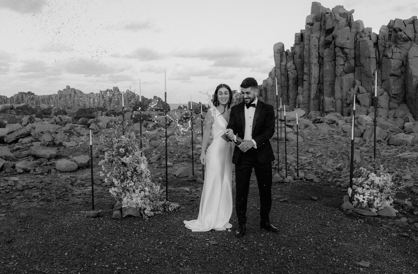 A couple in wedding attire stands before dramatic rock formations in a desert landscape with flower arrangements.