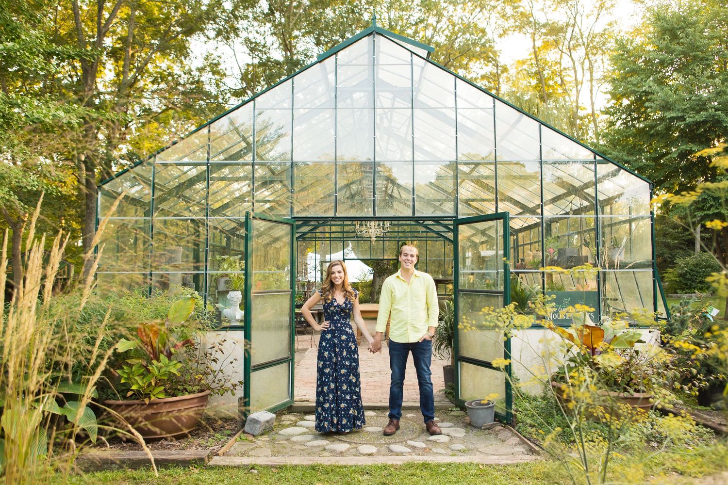 Engagement session with couple standing in front of glass greenhouse surrounded by lush garden.