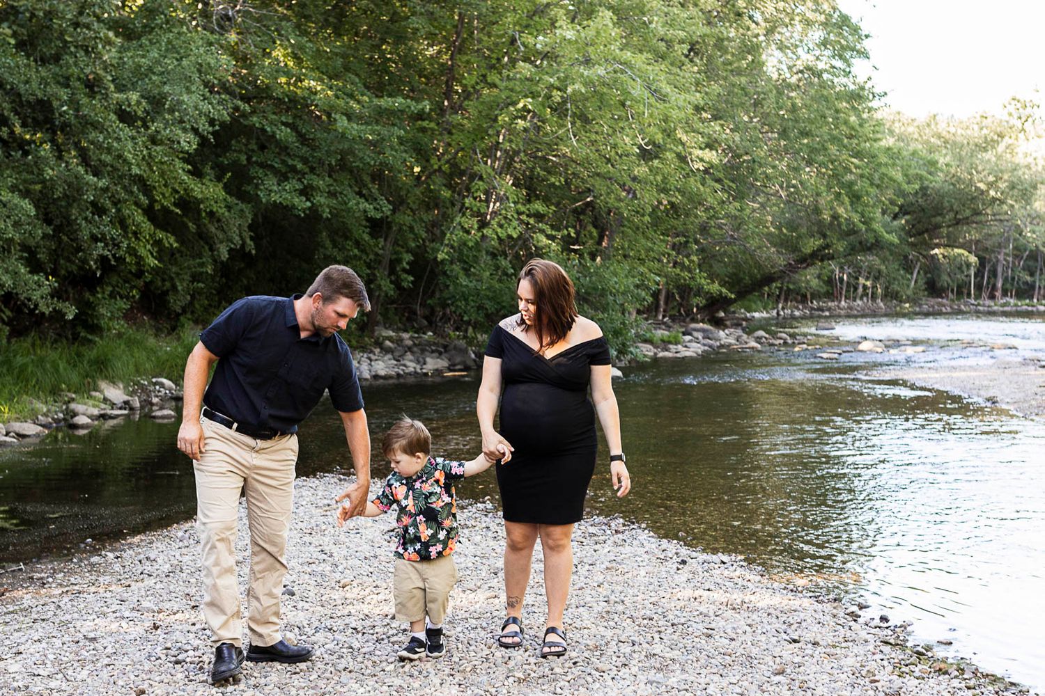 Pregnant mom, dad and little boy holding hands walking toward the camera. River and green trees behind them.