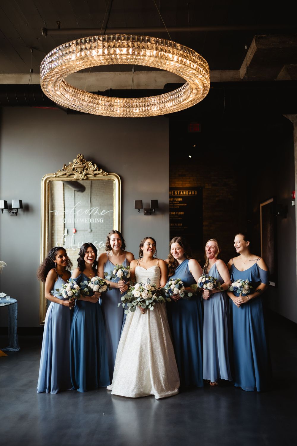 Wedding party in dusty blue dresses posed beneath ornate circular chandelier.