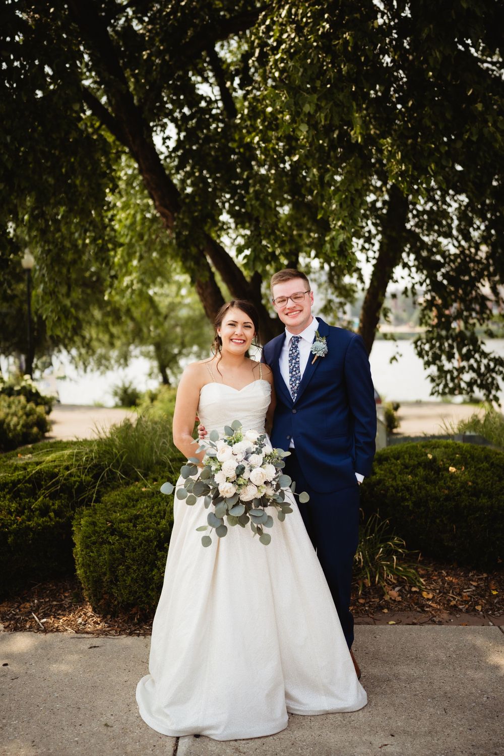 A wedding couple poses under a tree with the bride holding a white and green floral bouquet.