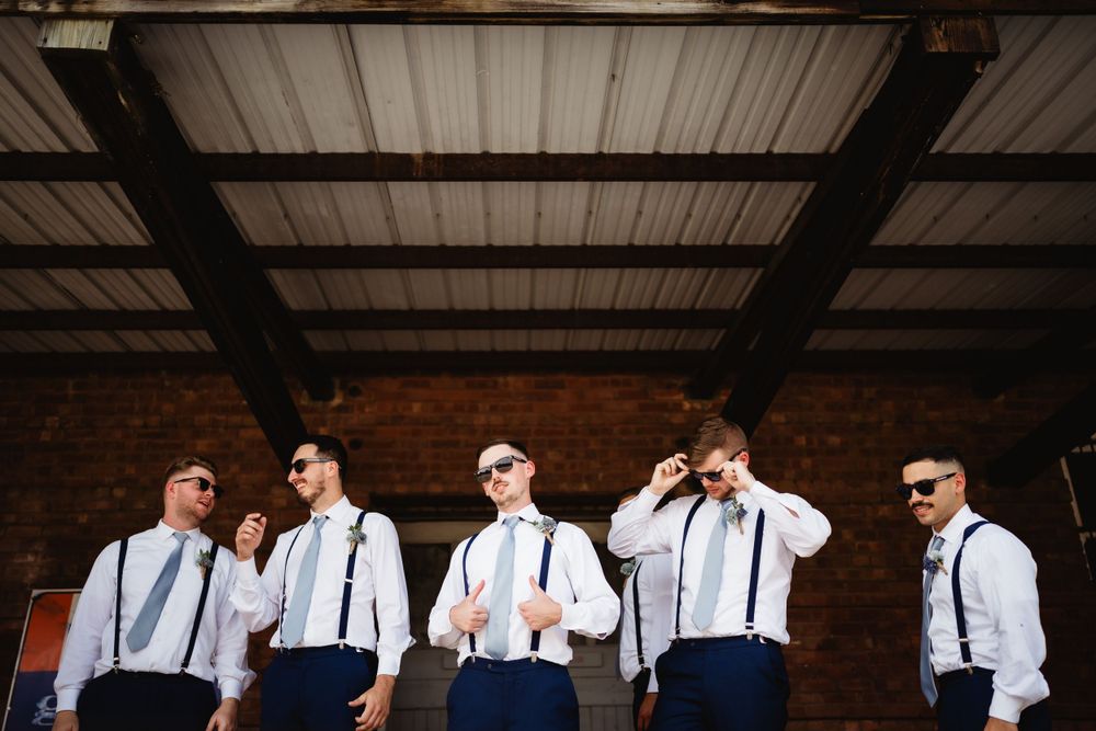 Groomsmen in navy pants and suspenders pose against industrial backdrop.
