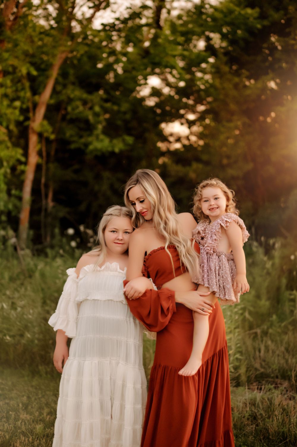 Three people in flowing dresses pose together in a sunlit meadow at golden hour.