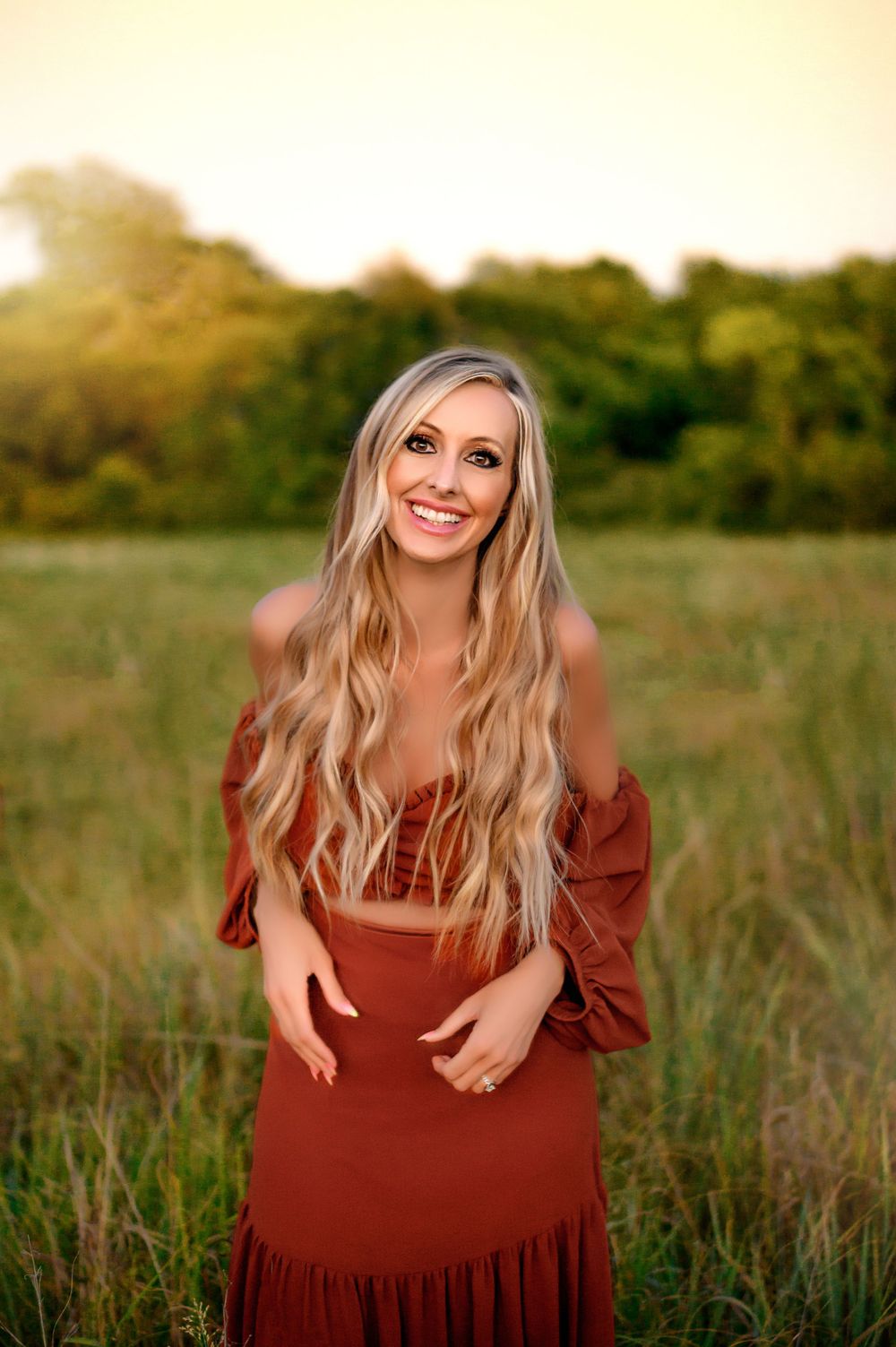 Woman in rust-colored dress poses in field during golden hour.