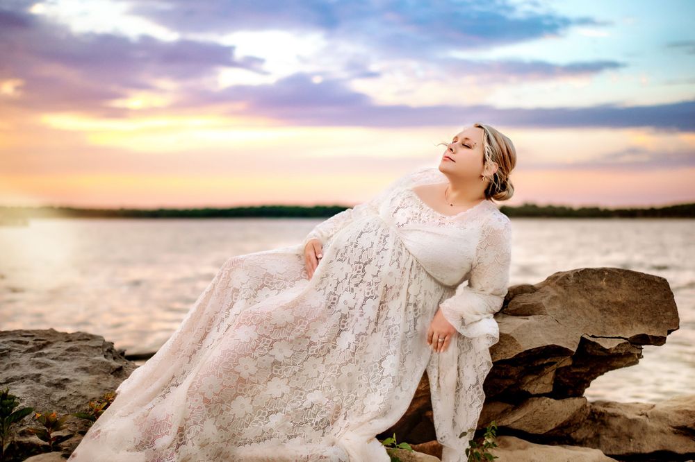 Person in lace dress poses dramatically on rocky shore at sunset.