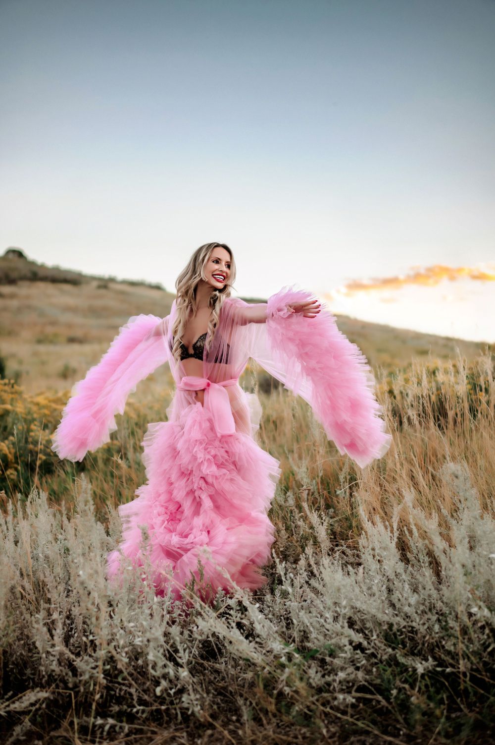 A woman in a flowing pink tulle dress poses dramatically in a field at sunset.