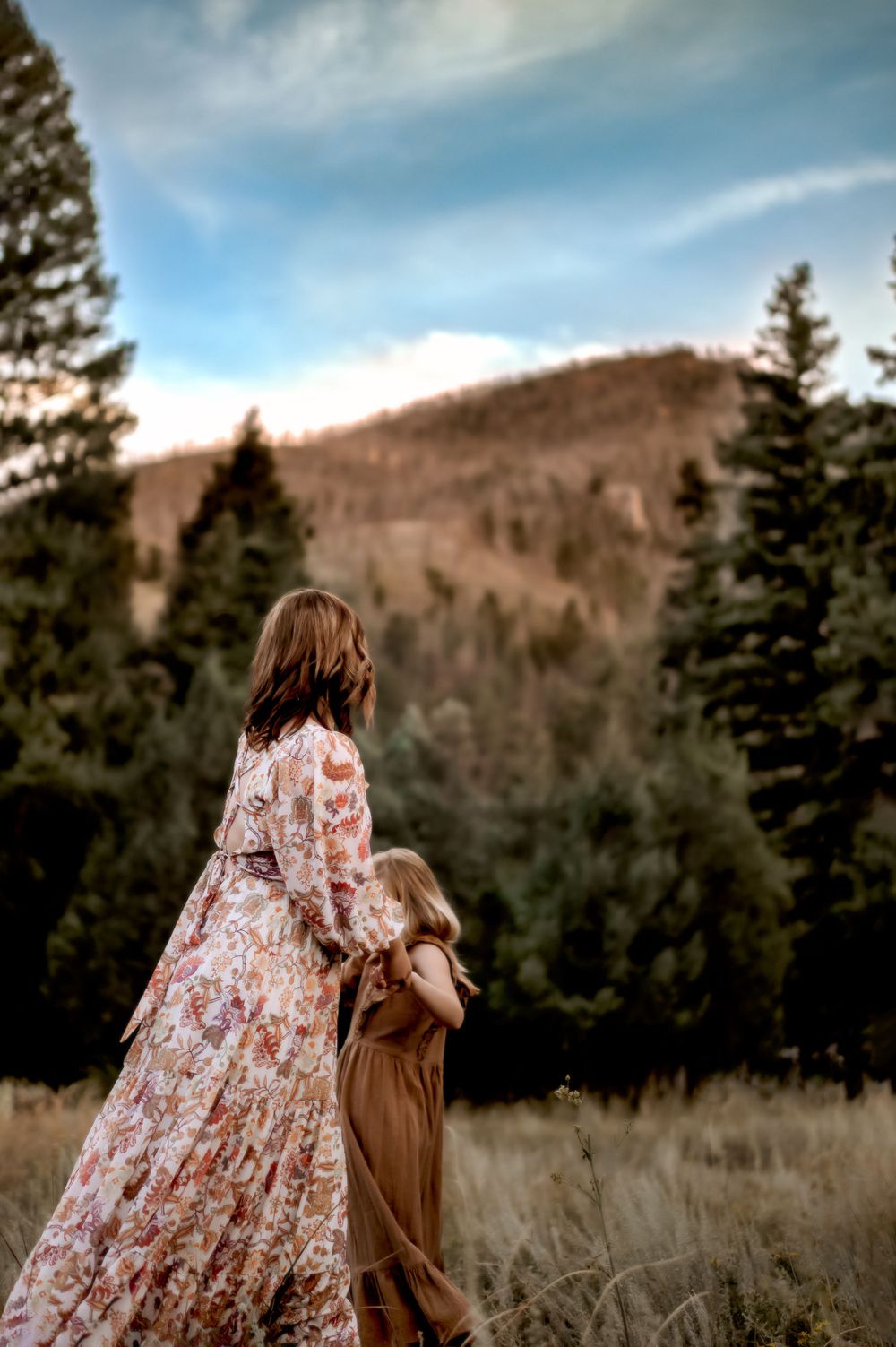 A floral dress and brown dress contrast against evergreen trees and mountain backdrop in nature.