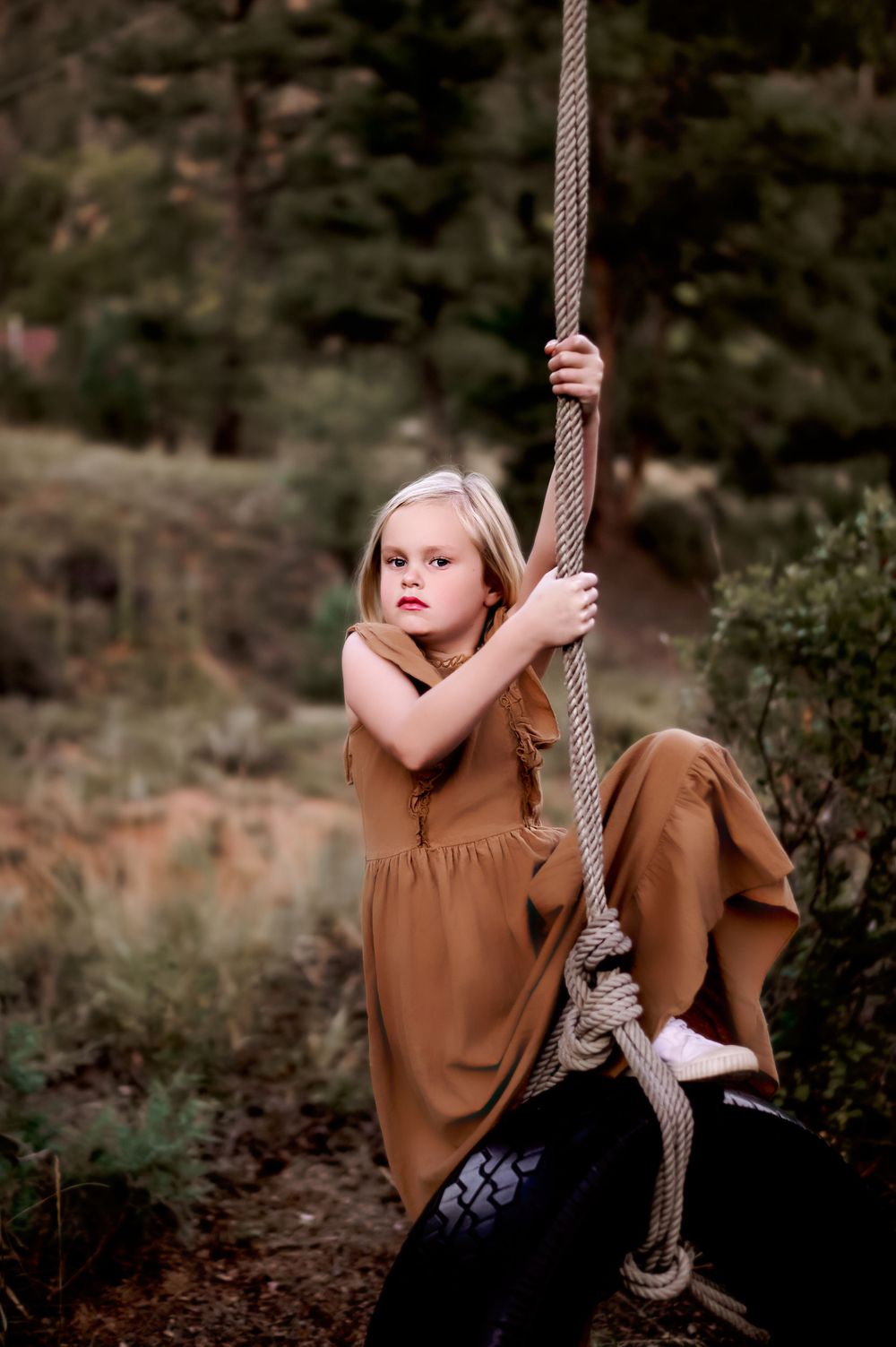 A young person in a brown dress sits on a tire swing in a rustic outdoor setting.