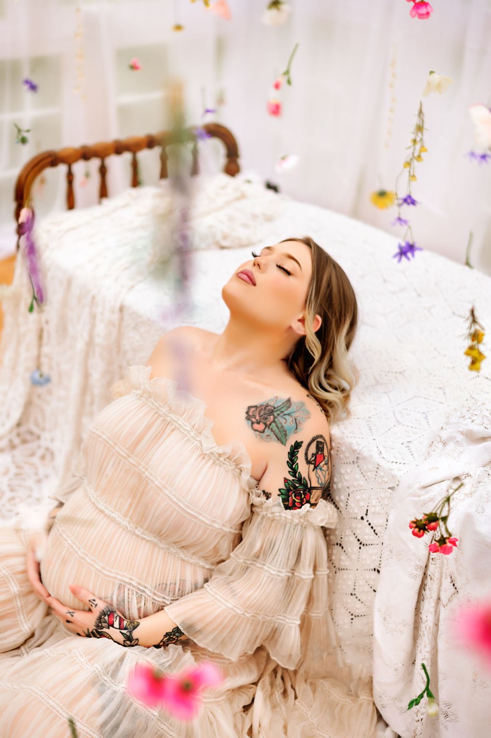 A romantic maternity photo featuring a cream-colored dress and delicate floral decorations hanging from above.
