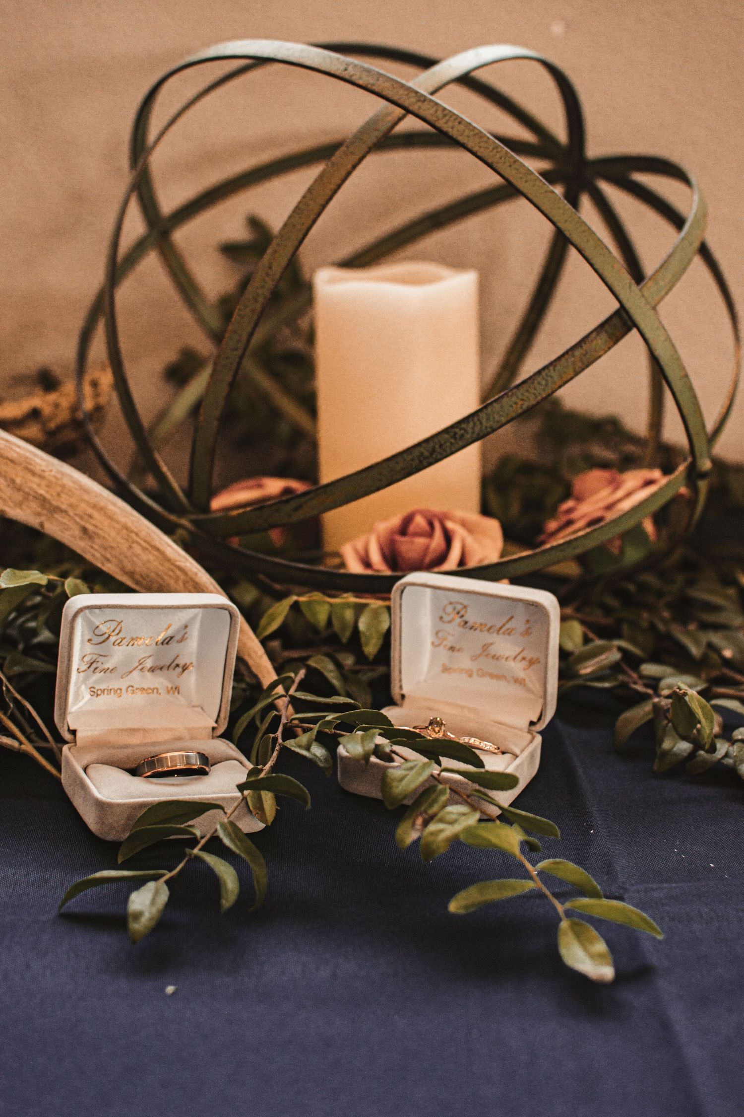 Wedding rings in vintage boxes displayed with a metal orb candle holder and roses on dark fabric.