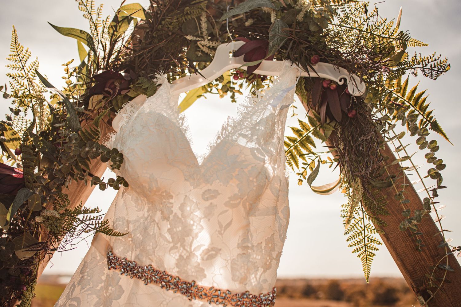 A delicate lace wedding dress hangs from a rustic arch decorated with ferns and greenery in golden sunlight.