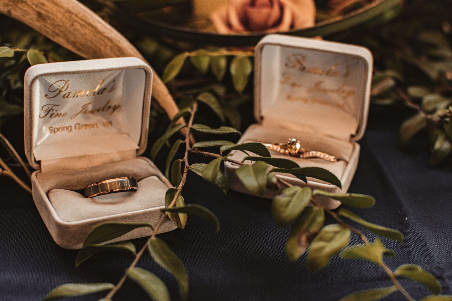 Two wedding ring boxes nestled among green foliage and roses in warm lighting.