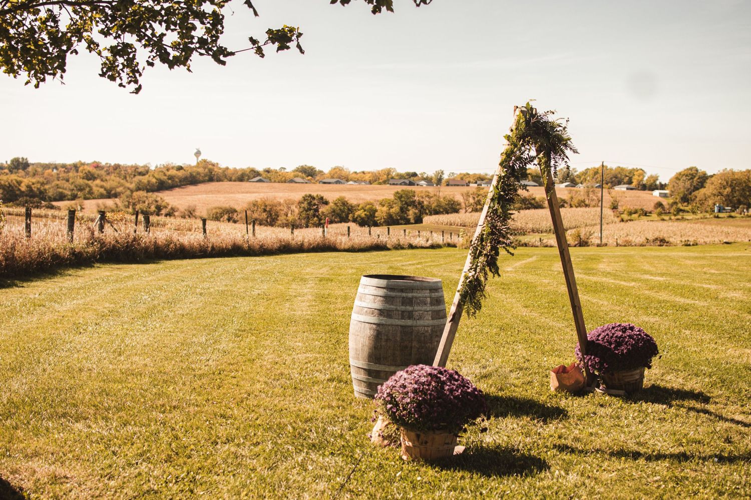 Rustic wedding arch decorated with greenery and flowers next to wine barrel in open field.