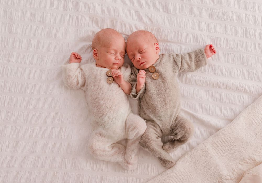 Twin newborns sleeping side by side in white and beige wraps on a textured background.