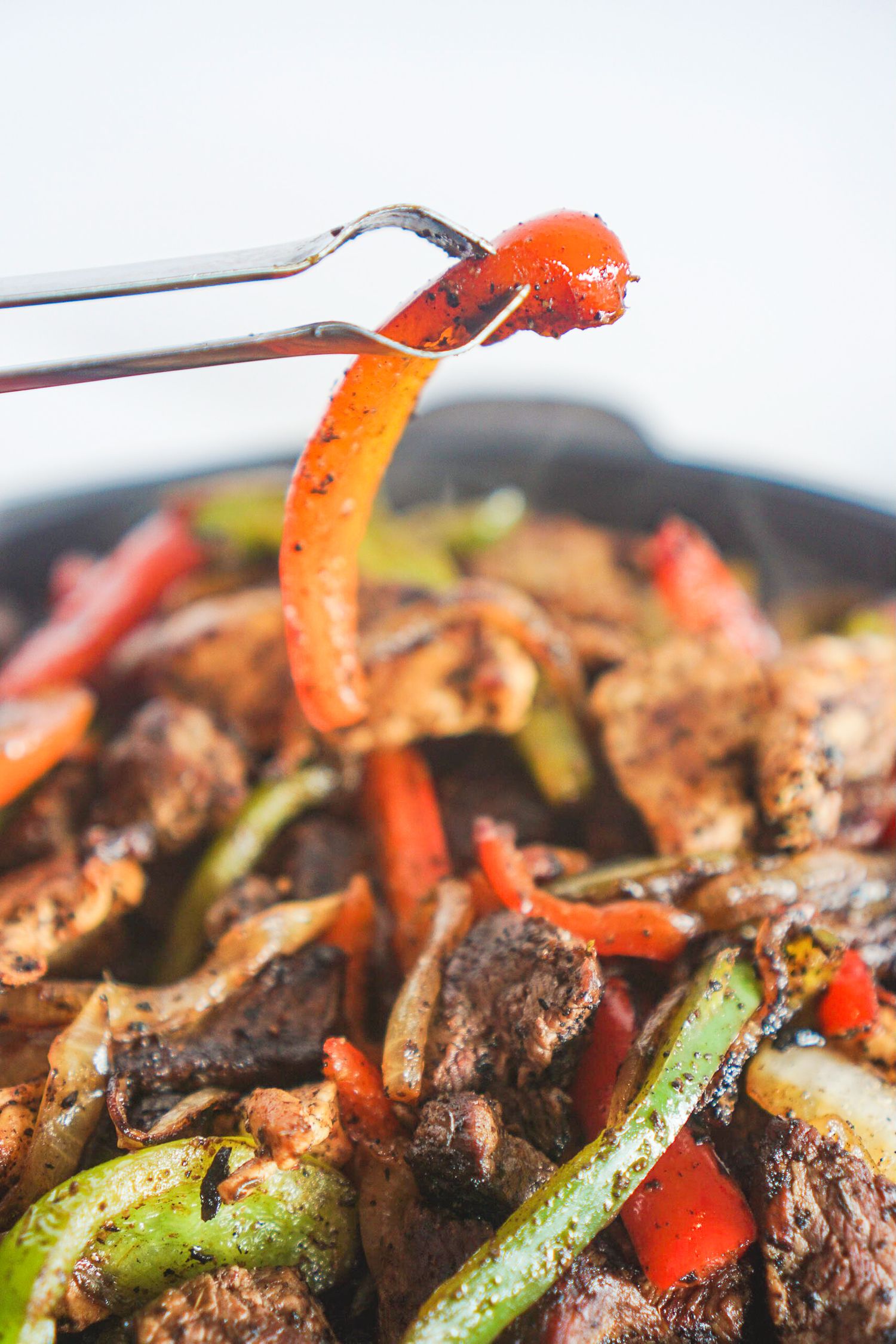 Red bell pepper strip held with tongs above a sizzling fajita dish