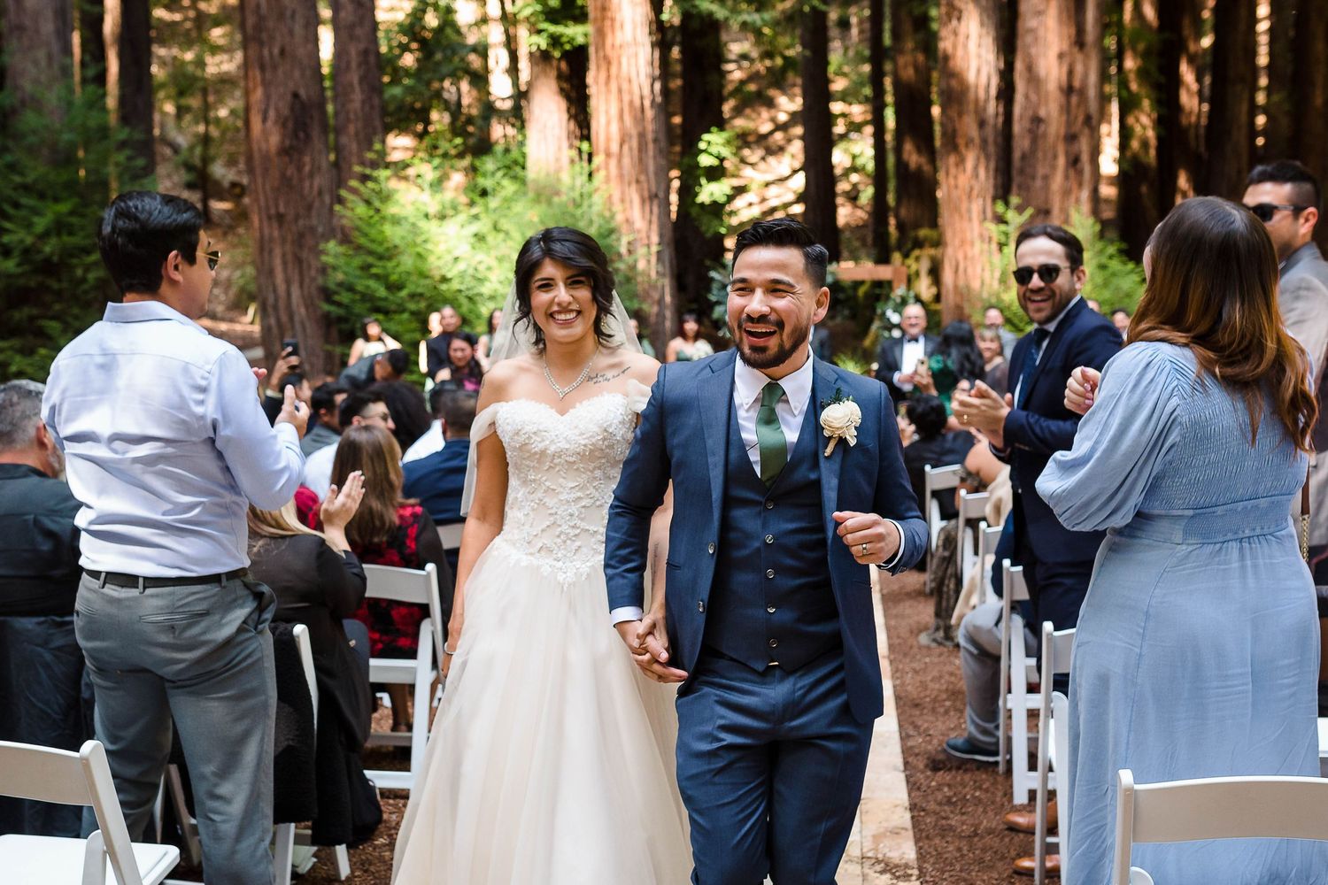 Newlyweds walk down the aisle in a redwood forest as guests celebrate.