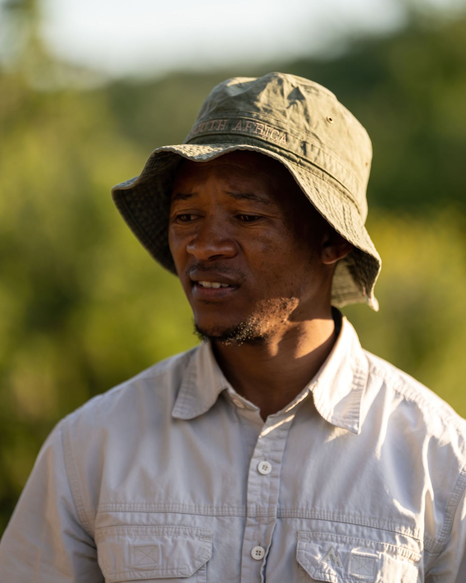 A person wearing a khaki hat and white shirt smiles warmly in natural outdoor lighting.