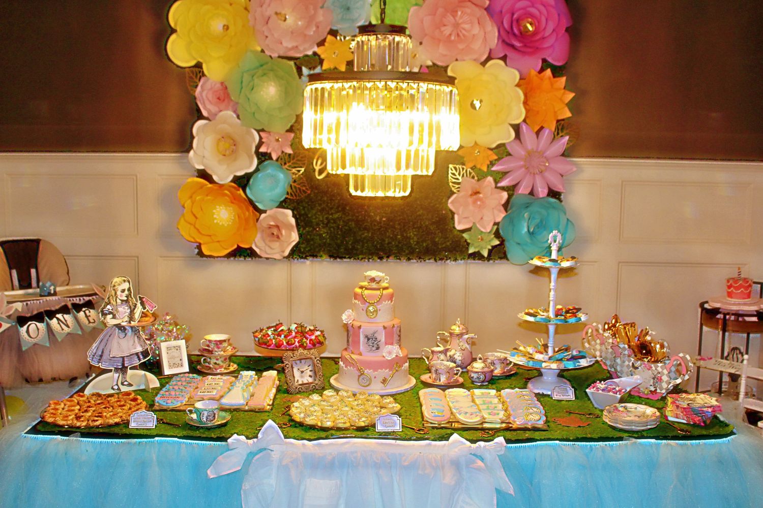 Colorful dessert table with chandelier and paper flower wall backdrop for special occasion.