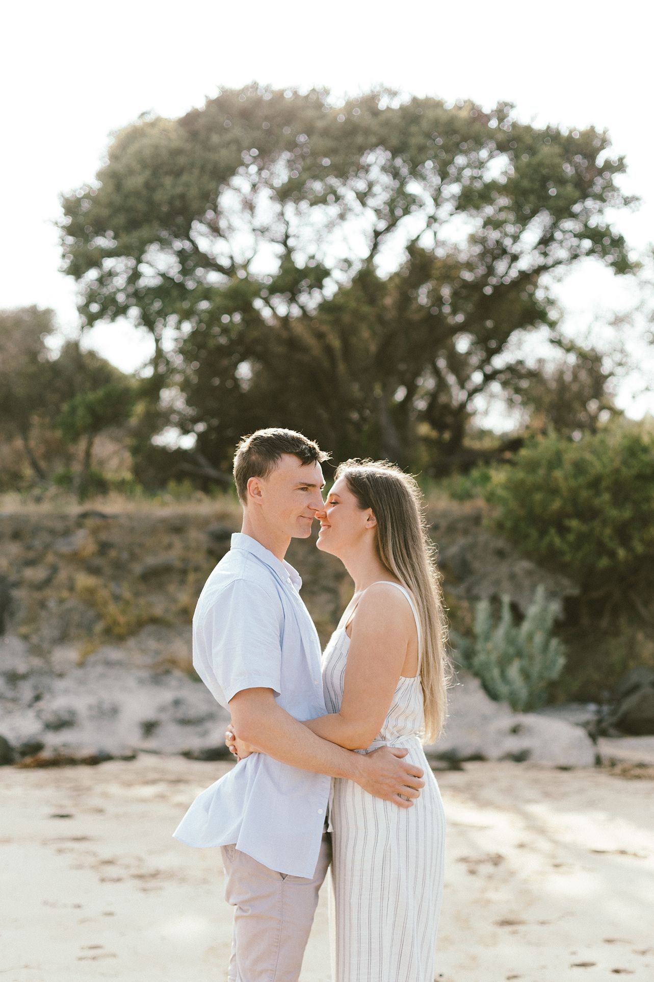 A romantic couple embraces on a beach with trees in the background during a sunset photoshoot.