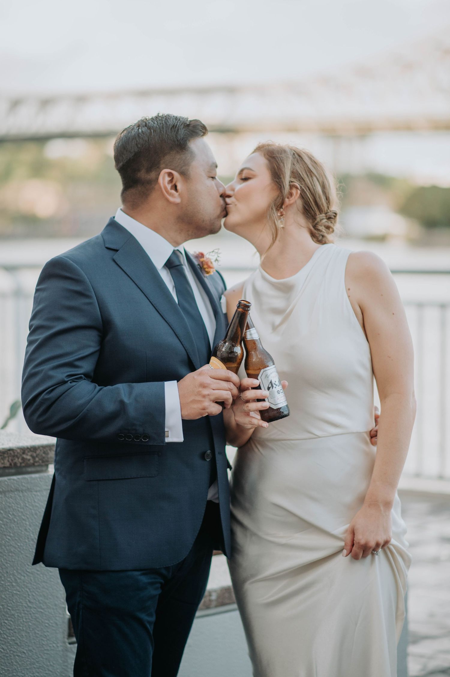 Newlyweds share an intimate kiss while holding beer bottles against a scenic waterfront backdrop.