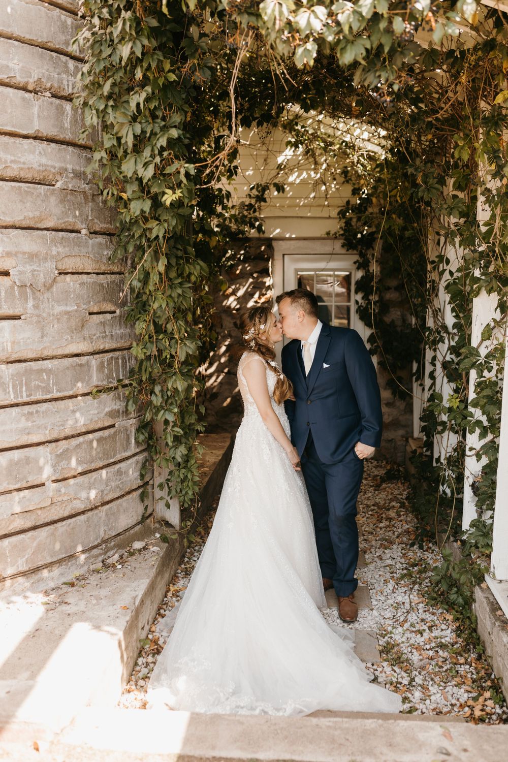 Bride and groom share a kiss under a lush green vine-covered archway in dappled sunlight.