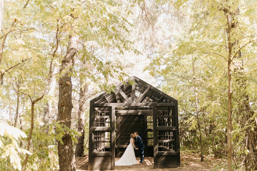 Bride and groom kiss at the entrance of a dark wooden greenhouse nestled among forest trees.