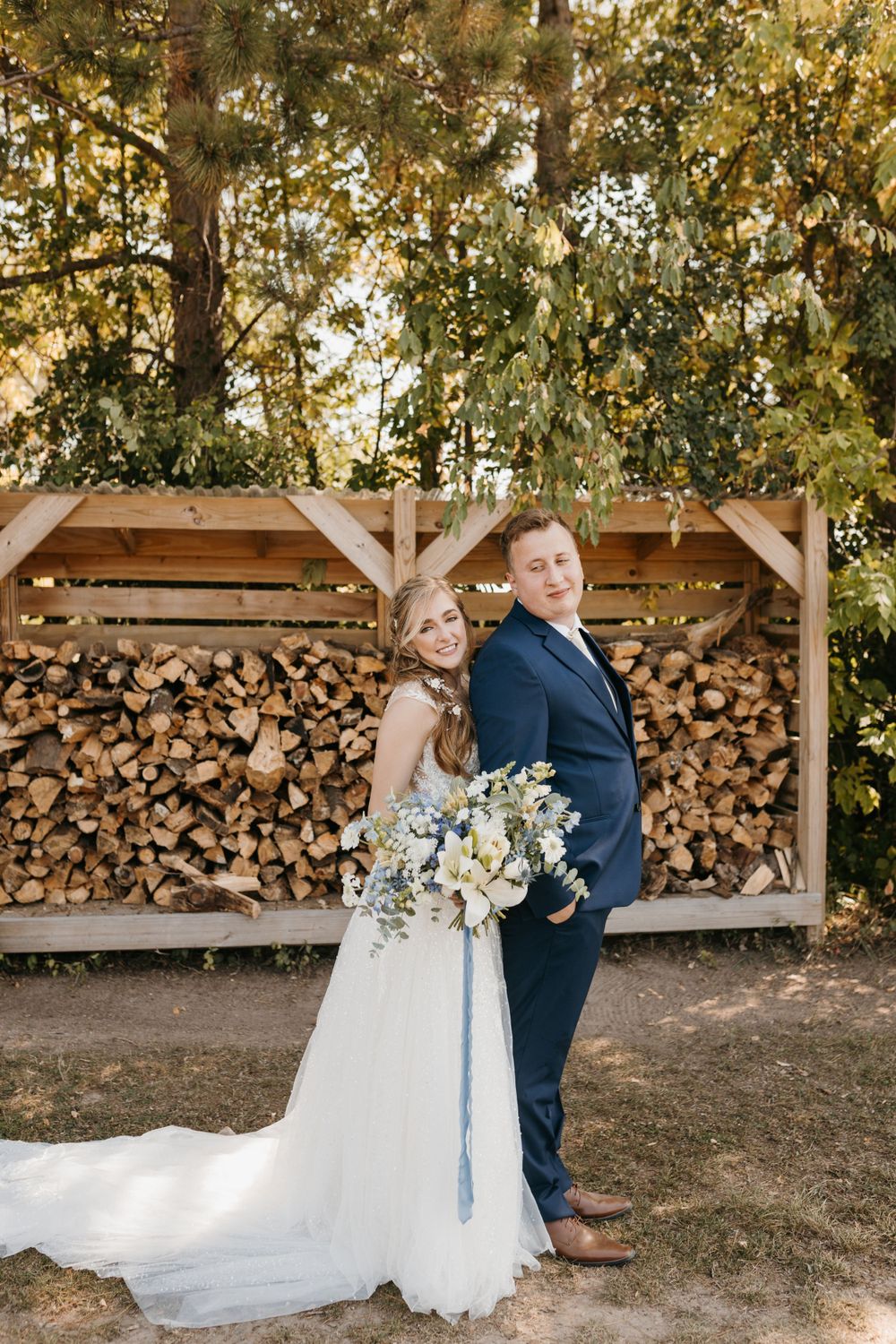 Bride and groom pose back-to-back in front of a rustic wood pile, bride holding a white and blue bouquet.