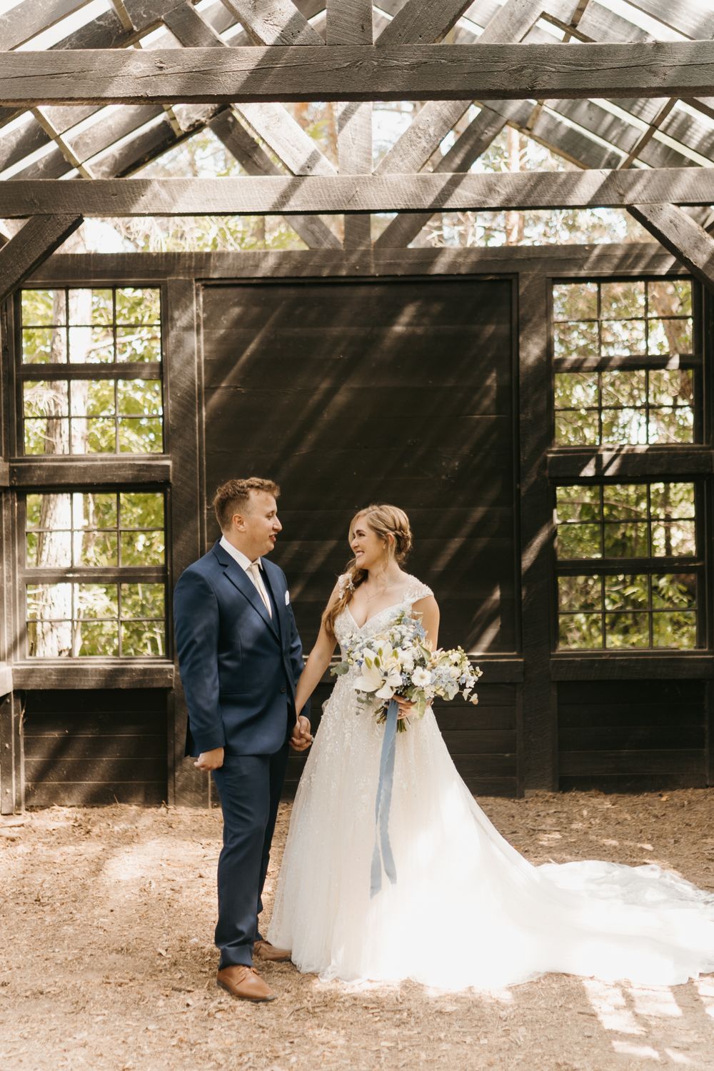 Bride with floral bouquet and groom hold hands gazing at each other outside a rustic dark wooden greenhouse.
