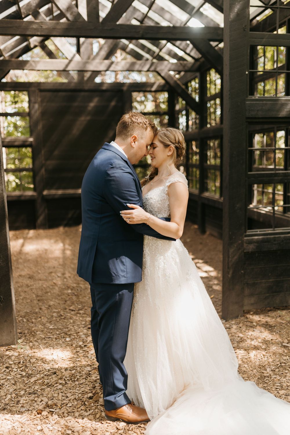 Bride and groom share a tender almost-kiss inside a rustic dark wooden greenhouse wedding venue in a forest.