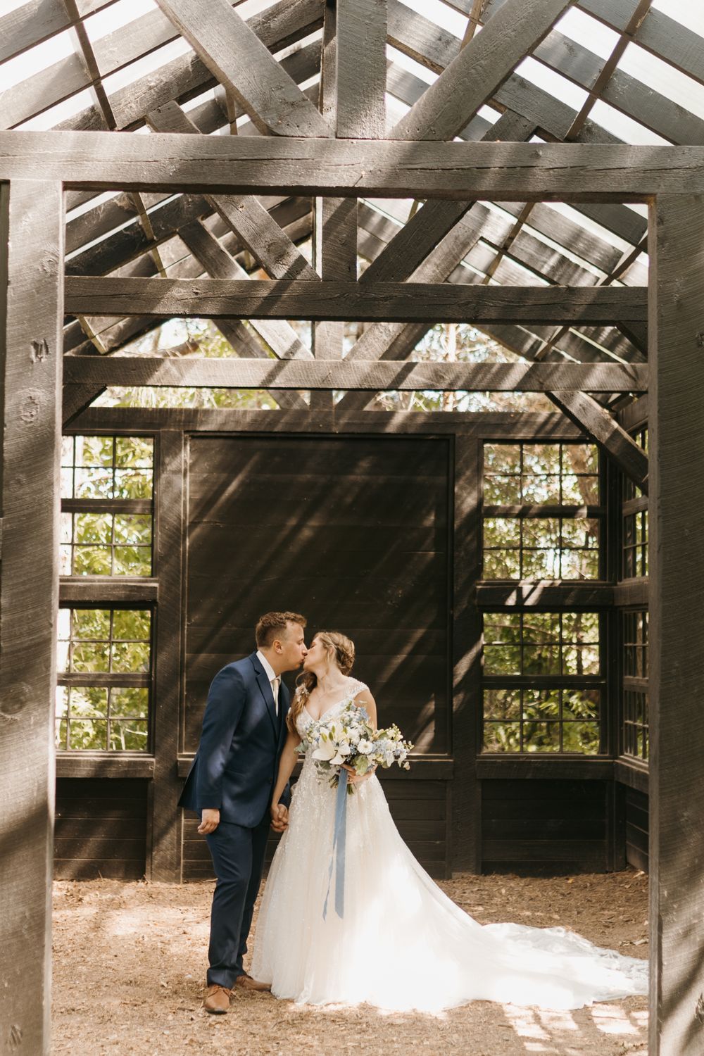 Bride and groom share a kiss inside a rustic wooden greenhouse structure surrounded by trees.