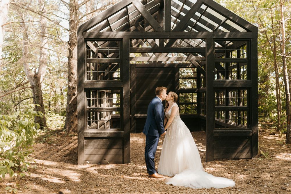 Bride and groom share a romantic kiss in front of a dark wooden greenhouse structure surrounded by trees.