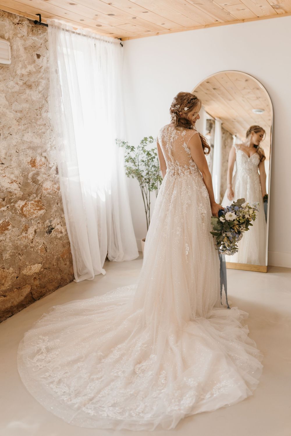 Bride in lace wedding gown holds blue floral bouquet while gazing into arched mirror in a sunlit rustic bridal suite.