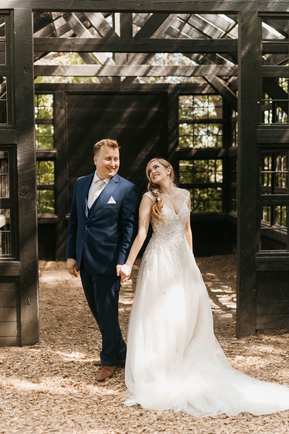 Smiling bride and groom hold hands walking together in front of a rustic dark wood greenhouse structure.