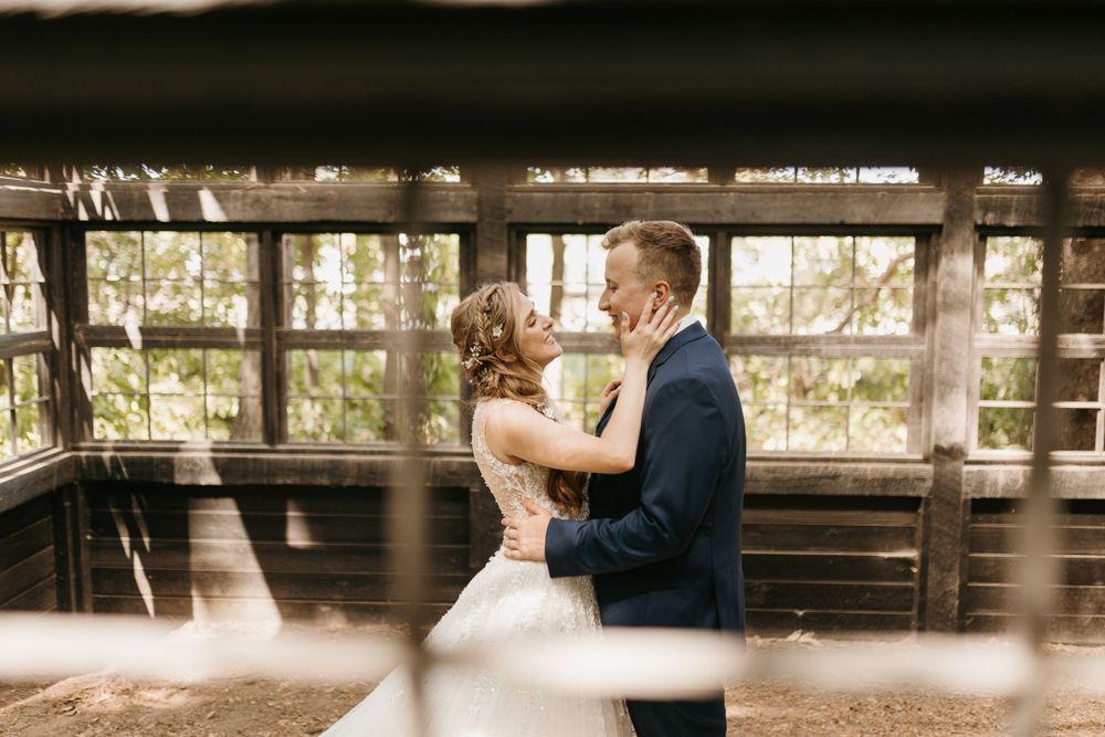 Bride and groom share an intimate moment inside a rustic wooden structure with large windows.