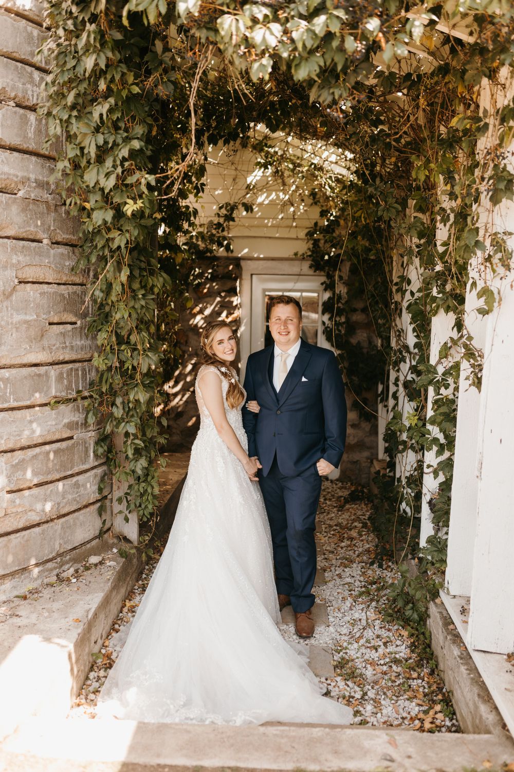 Bride and groom walk hand-in-hand through a vine-covered archway at an outdoor wedding venue.