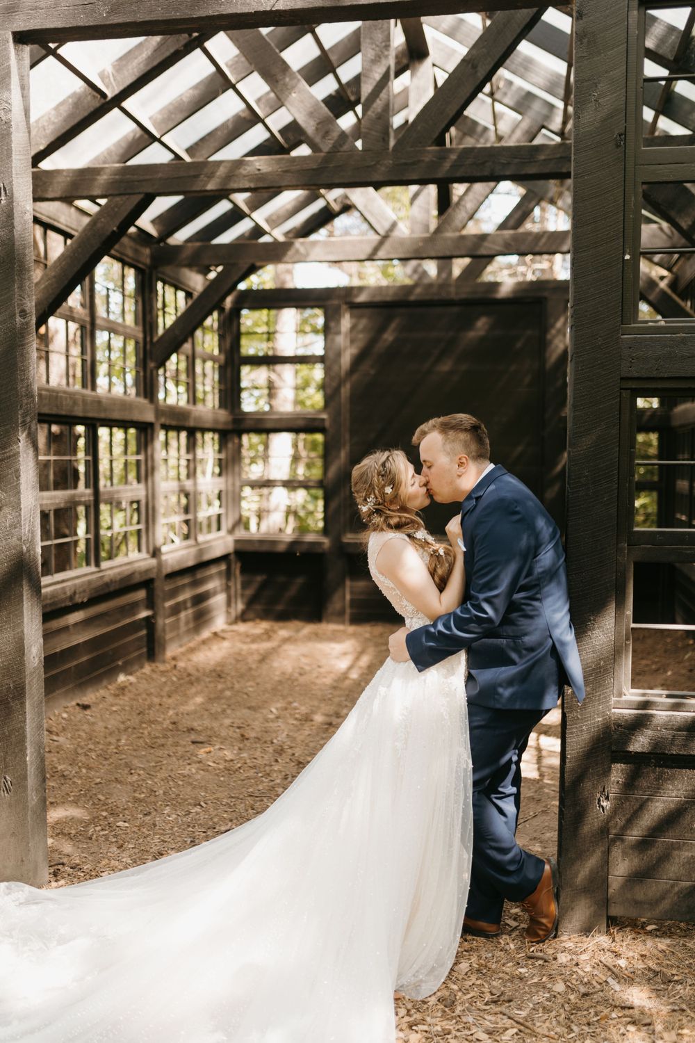 Wedding couple shares a romantic kiss inside a rustic wooden greenhouse with large windows.