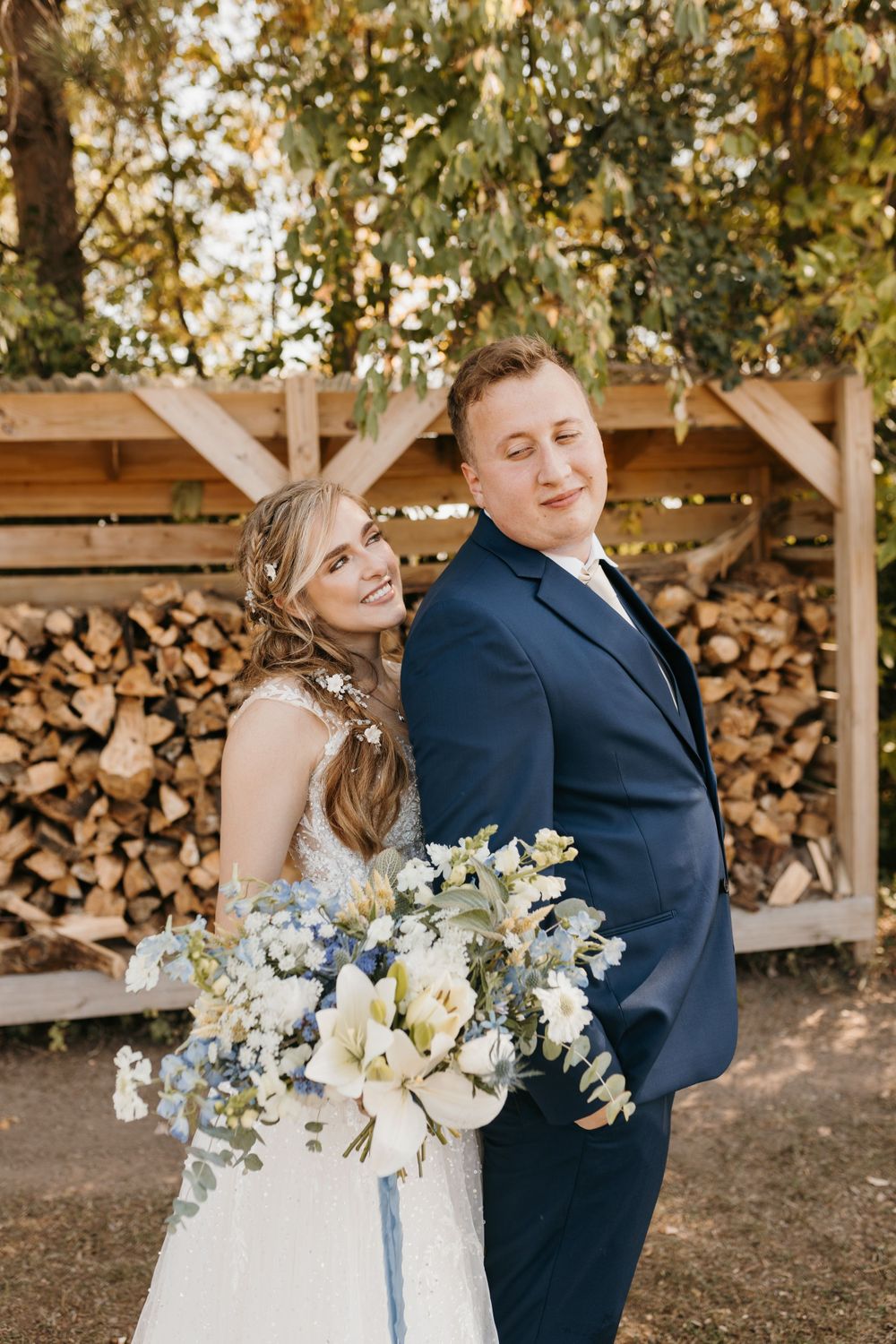 Smiling bride holds a white and blue floral bouquet while standing back-to-back with groom in navy suit.