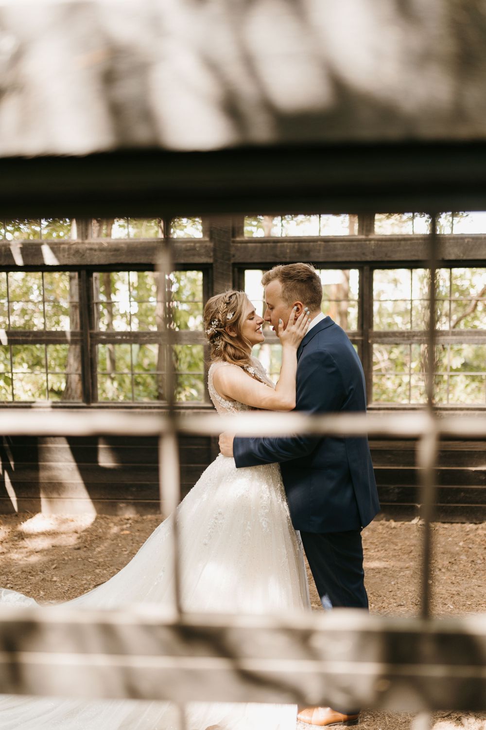 Bride and groom nearly kiss inside a rustic wooden greenhouse with bright windows behind them.