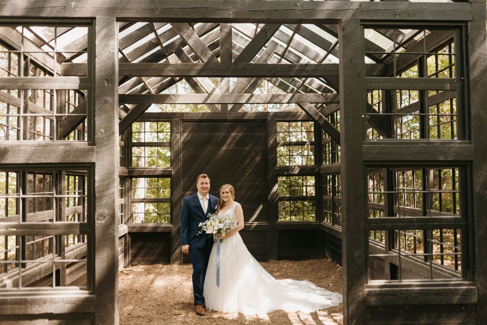 Bride and groom pose together inside a stunning rustic wooden greenhouse wedding venue surrounded by tall trees.